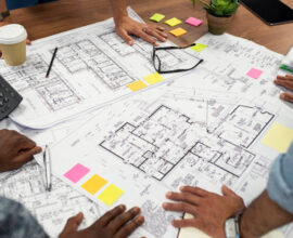 Closeup of multiethnic hands on blueprints at architects office. Team of designer and engineers working together on new residence complex. Top view of blue prints layout of house and buildings on wooden table.